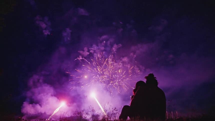 Colorful fireworks bursting and exploding in the sky reveal the silhouette of a couple sitting in the grass