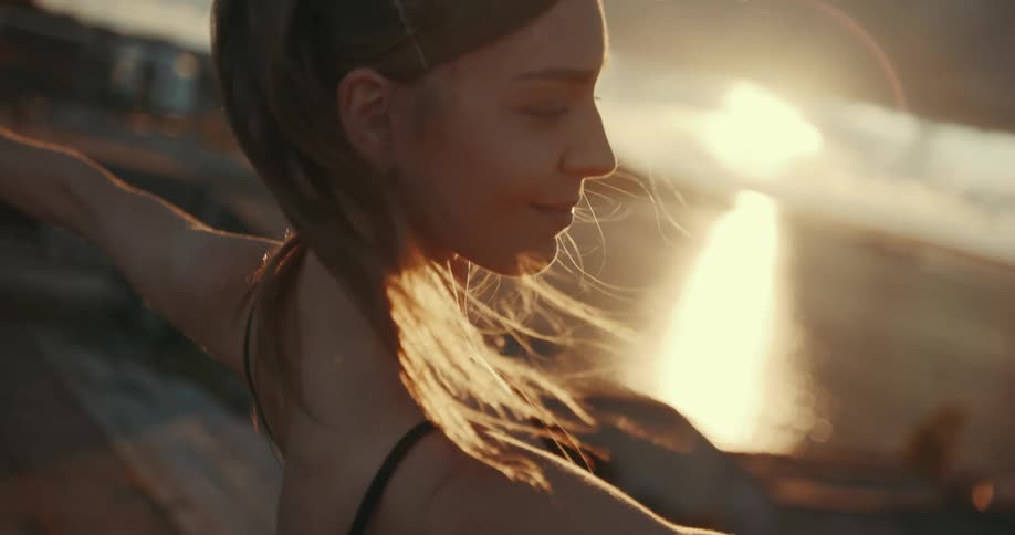 A woman smiles softly as she leans back into a dance pose stretch with sunlight shimmering on the water surface