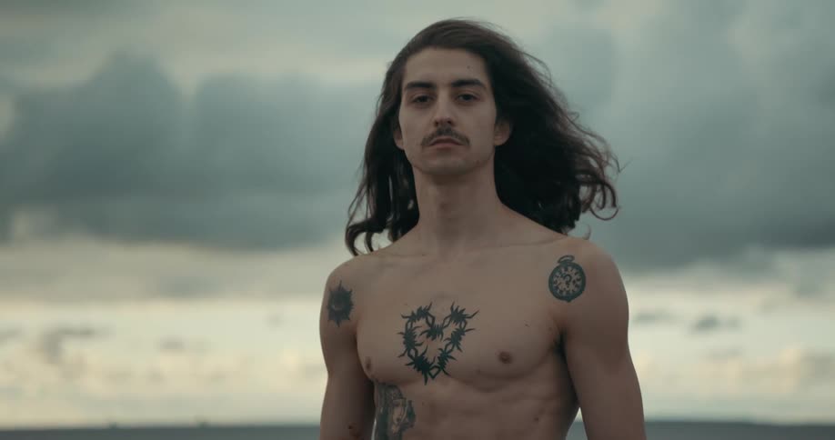 A tattooed man stands outside topless