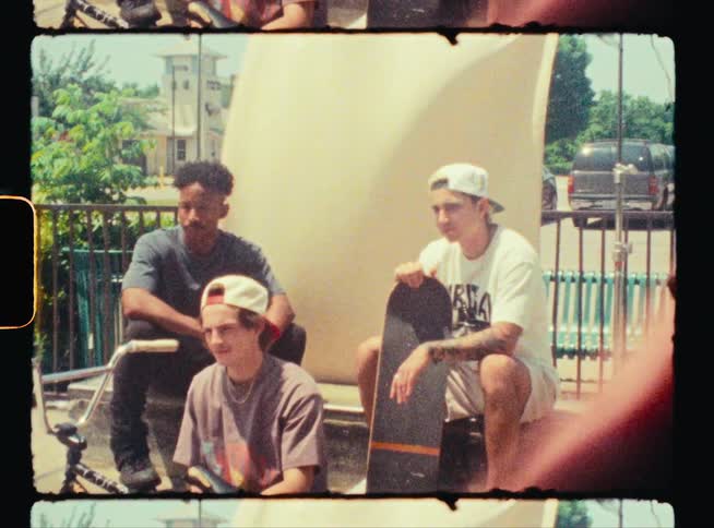 Three teenage boys are sitting together in a park and one is behind a bike and the another is holding a skateboard