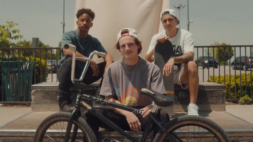 Three teenagers sit and pose for a photo one holding a skateboard and one holding his bike