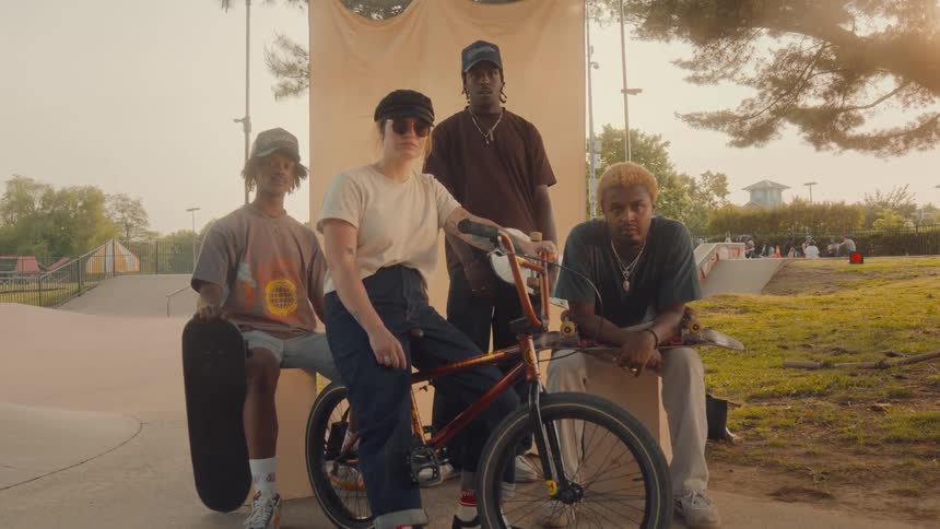 A group of teenagers one on a bike and the others holding skateboards pose for a photo by a park