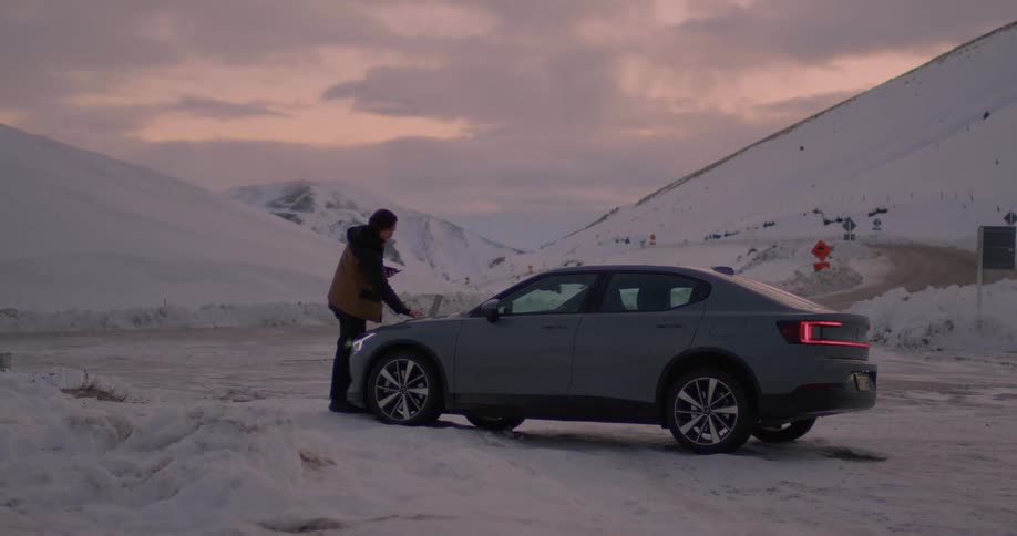 A man is parked on a snow covered mountain road and standing outside typing on a laptop computer on the hood of his car as he holds his cell phone