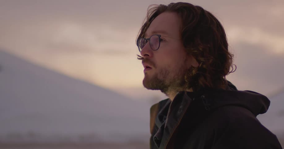 Young long haired bearded man wearing glasses sitting on the ground at winter working on a laptop