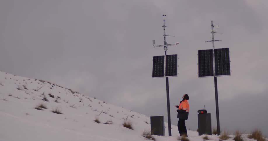 A man is standing outside holding a laptop computer and looking up at utility poles with solar panels