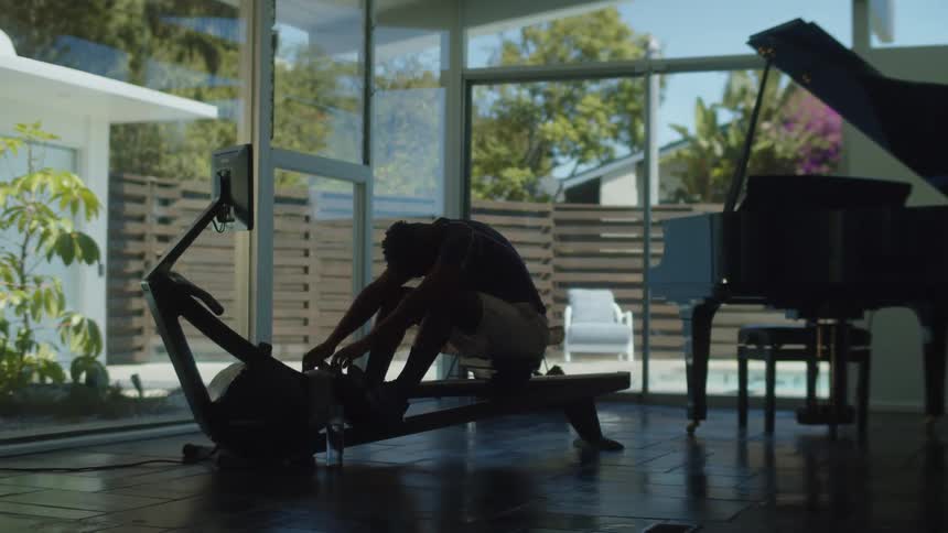 A man sitting on a rowing machine wipes the sweat off his face as he looks out at his patio