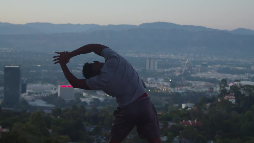 A man does stretches on a hill overlooking a city during the day