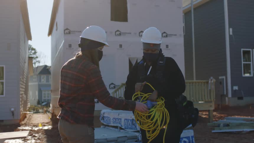 Two construction workers work with power cord on construction site as vehicles pass in the background