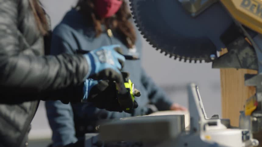One woman watches as another uses a tape measurer at workbench with a table saw