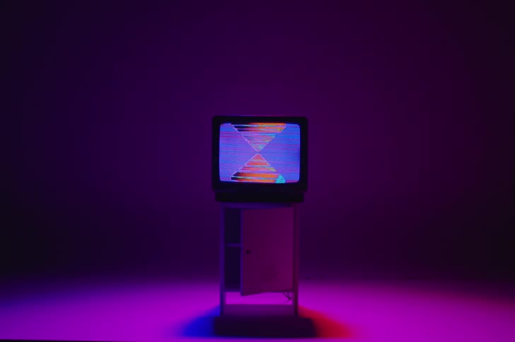 Square and triangle shapes in purple and blue static move on a television screen sitting on a floor illuminated with a magenta colored light