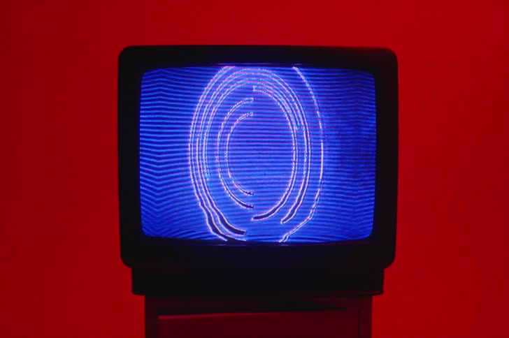 A circular pattern and designs move and flash on a television screen sitting in a deep red room