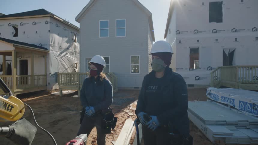 Construction workers stand near power tools as sun shines over homes under construction in the background