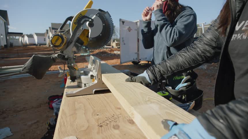 A woman holding a board hands another woman a pair of safety goggles and she puts them on and prepares to saw the wood