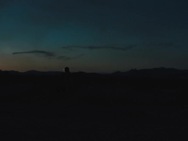 Person wearing headlamp walks across dark ground and is silhouetted against slightly red tinted horizon