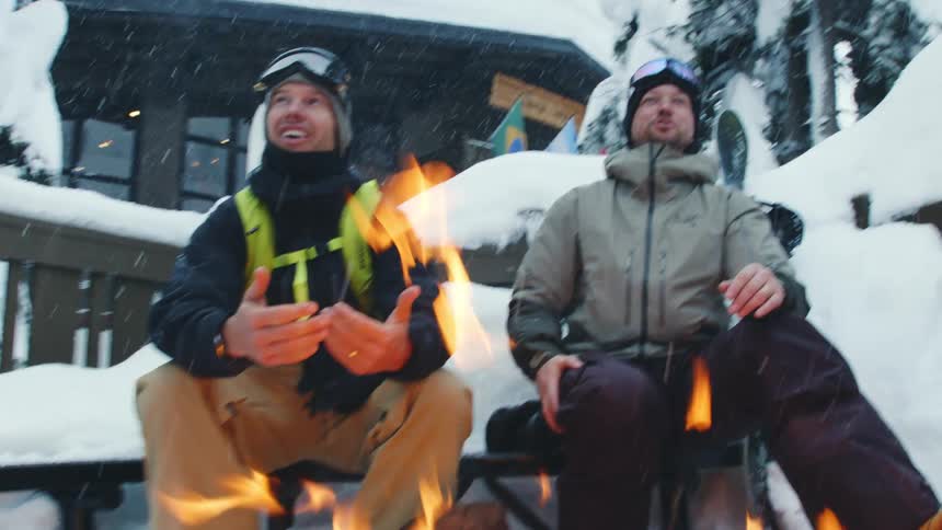 Two men laugh while warming their hands over a bonfire outside a lodge
