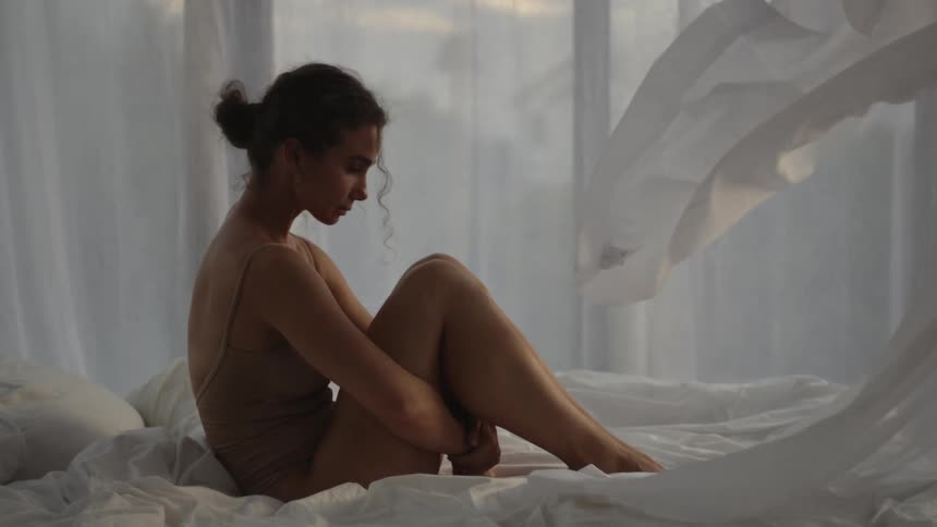 Curtains surrounding a bed drape over a woman hugging her thighs while sitting atop the bed