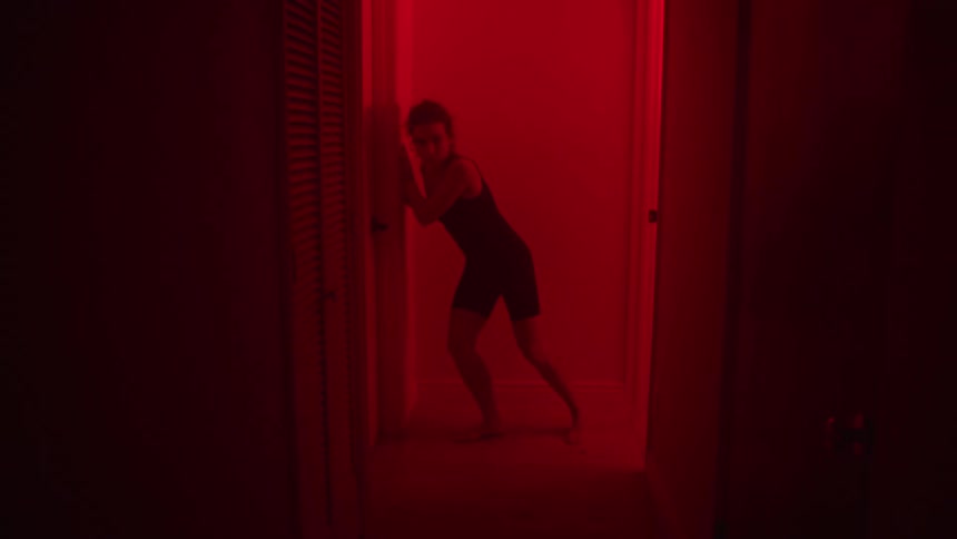 A woman hits a wall then runs through an interior corridor all lit by a reddish light