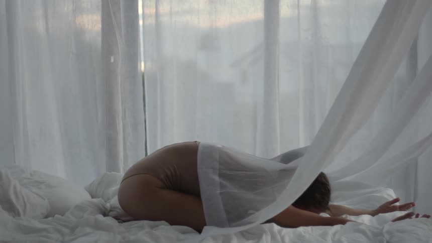 A woman lies in the fetal position in bed as curtains billow around her