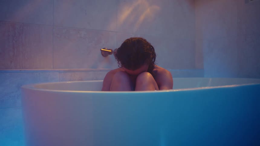 A woman sits in a bath tub with her face buried in her knees