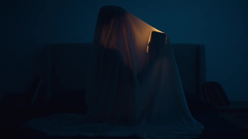 A person sitting beneath a sheet holds up a small light which they turn on