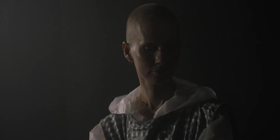 Bald woman standing in elaborate fashion against dark background looks down