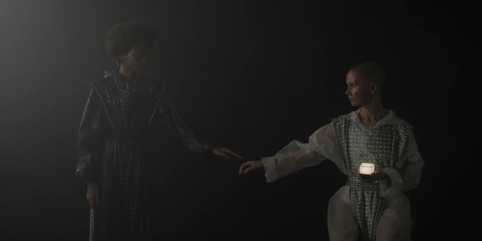 A teenage girl sits in the dark holding a light and a teenage boy stands beside her and they reach their arms out to touch hands