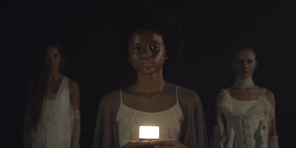 A black teenager holding a light stands outside in the dark in front of two other teenagers and they all just stare straight ahead