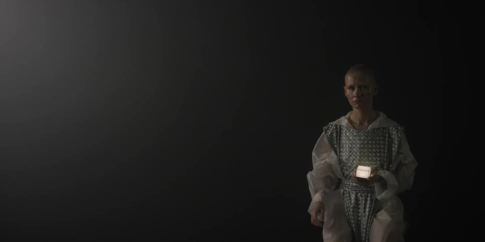A teenage girl sits in the dark holding a small light lowers her arm to her side and gets up and walks away