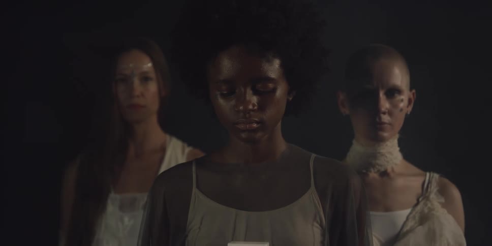 Three young woman stand in the darkness as one holds a light and lowers head to look at the light