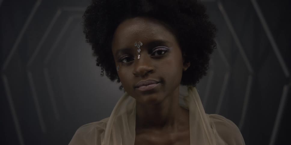 A woman wearing a sheer top and jewelry on her forehead looks up and smiles softly