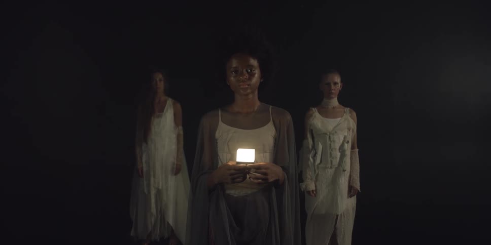 Three women are standing together in the darkness and the woman in front is holding a glowing cube