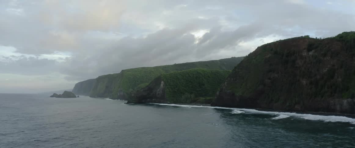 Ocean waves splash and foam against the shore lined with green cliffs and ridge