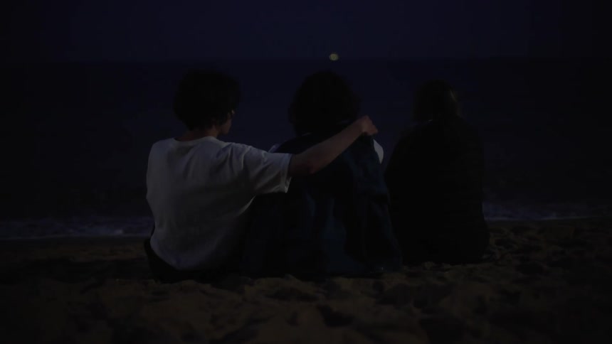 A person wrapping a sweater around their friends shoulders while sitting on a sandy beach at night