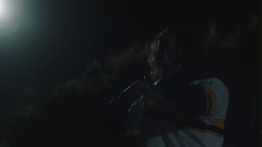 A man is standing outside in the darkness as a bright light shines from above and he pulls his shirt off over his head