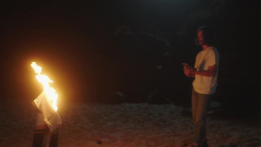 Man holding smartphone on beach at night sets fabric on fire with a lighter