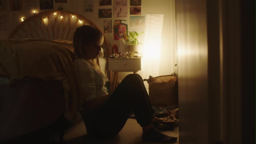 Woman sits on the floor and leans against the bed in dark room