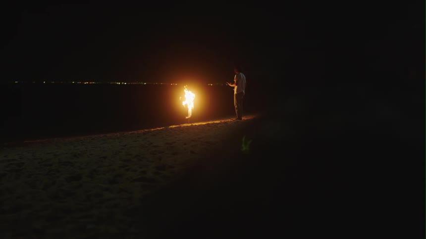 Young man lights up a fire on the white sand beach at night next to the ocean