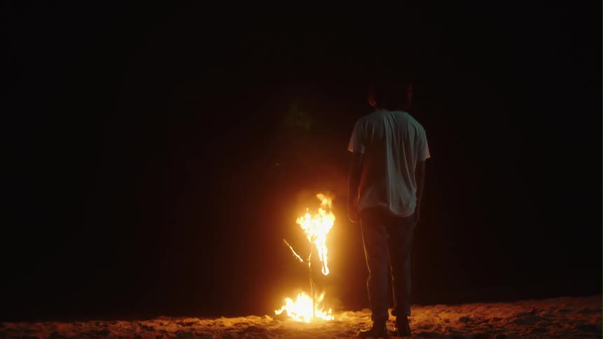Young man lights up a fire on the white sand beach at night next to the ocean