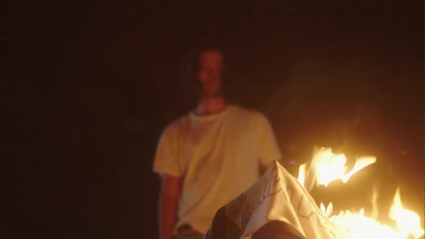 Man with long hair beard and white t shirt puts phone in pocket and watches bonfire on the beach at night