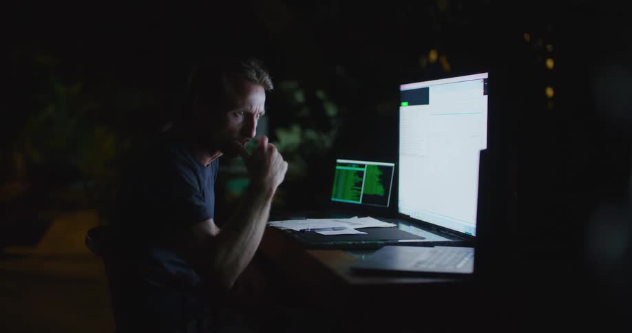 A man is sitting at a desk with a large monitor in front of him and a laptop on each side as he types on the keyboard to the right and looks back and forth at the screens
