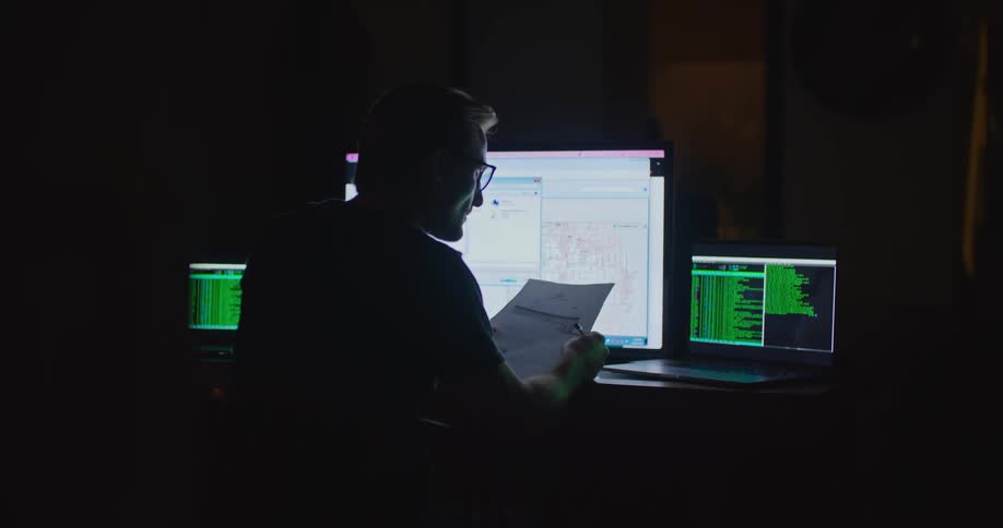 A man sits in front of computers in an office and twirls a pen as he looks at a paper and the monitors