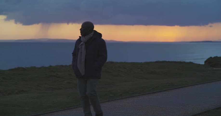 A man is walking on a costal path as clouds hang over the water