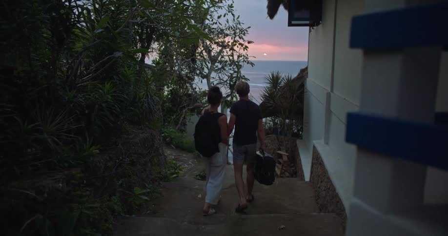 Middle aged blonde man wearing dark blue shirt and shorts holds womans hand wearing backpack and white pants go down the stairs next to buildings in town at sunset