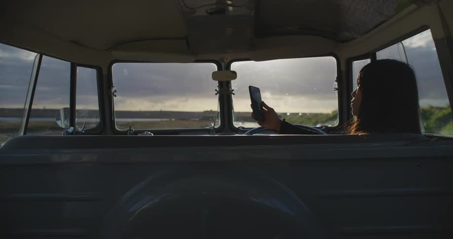 A woman stretches her legs out and up onto the dash in a classic van as she scrolls on her phone in her van