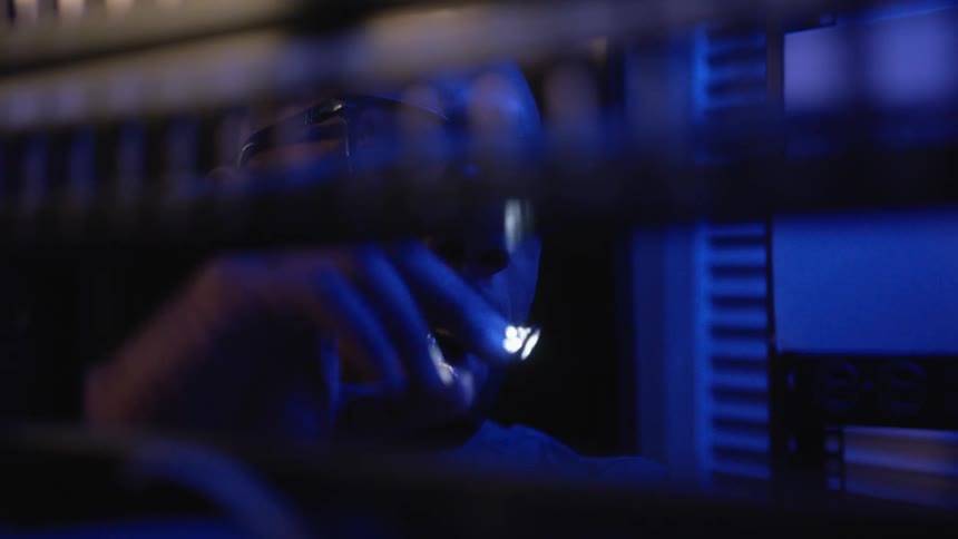 A man wearing glasses is in a dark room and he is holding a small flashlight in his mouth as he looks at items near him