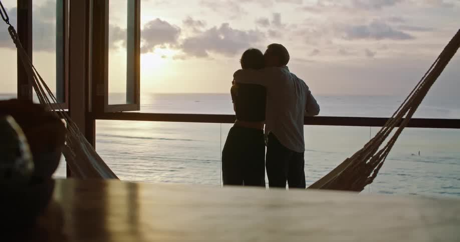 A man and woman stand on a deck on the shore drinking wine and he puts his arm around her and kisses her