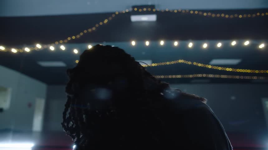 A person with dreadlocks is standing in a room with strings of lights across the ceiling rotating their head and their hair is moving in motion as they put each hand on their head