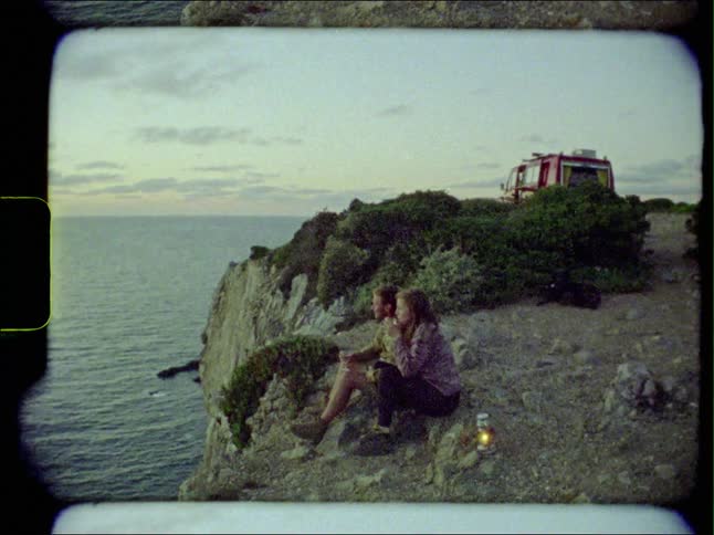 Young couple sitting on the edge of a cliff drinking next to the ocean at sunset