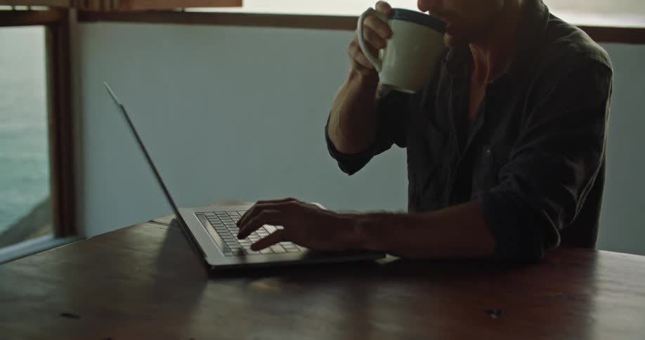 A man sits at a table drinking coffee and typing on a laptop