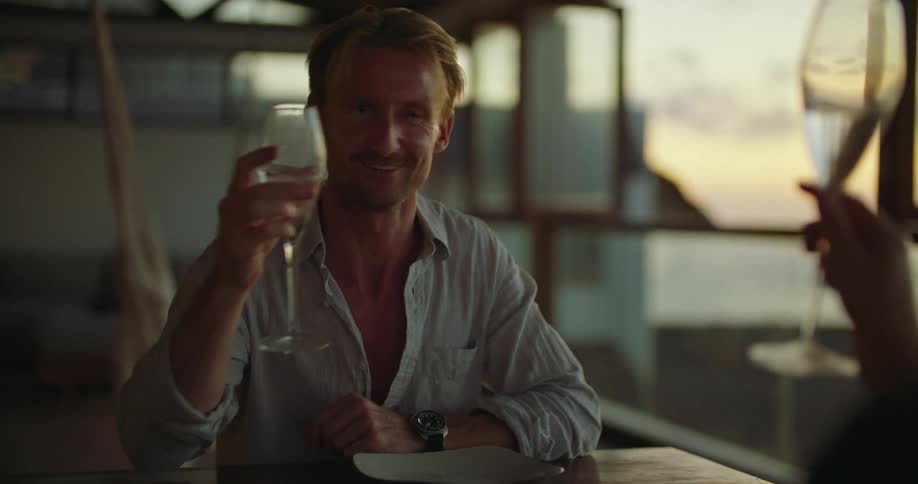 Middle aged blonde man with blue shirt and black wristwatch clinks glasses with woman sitting at table drinking wine
